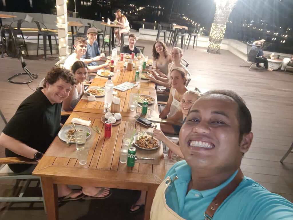 A man takes a selfie with efterskole students in the background sitting around a table 