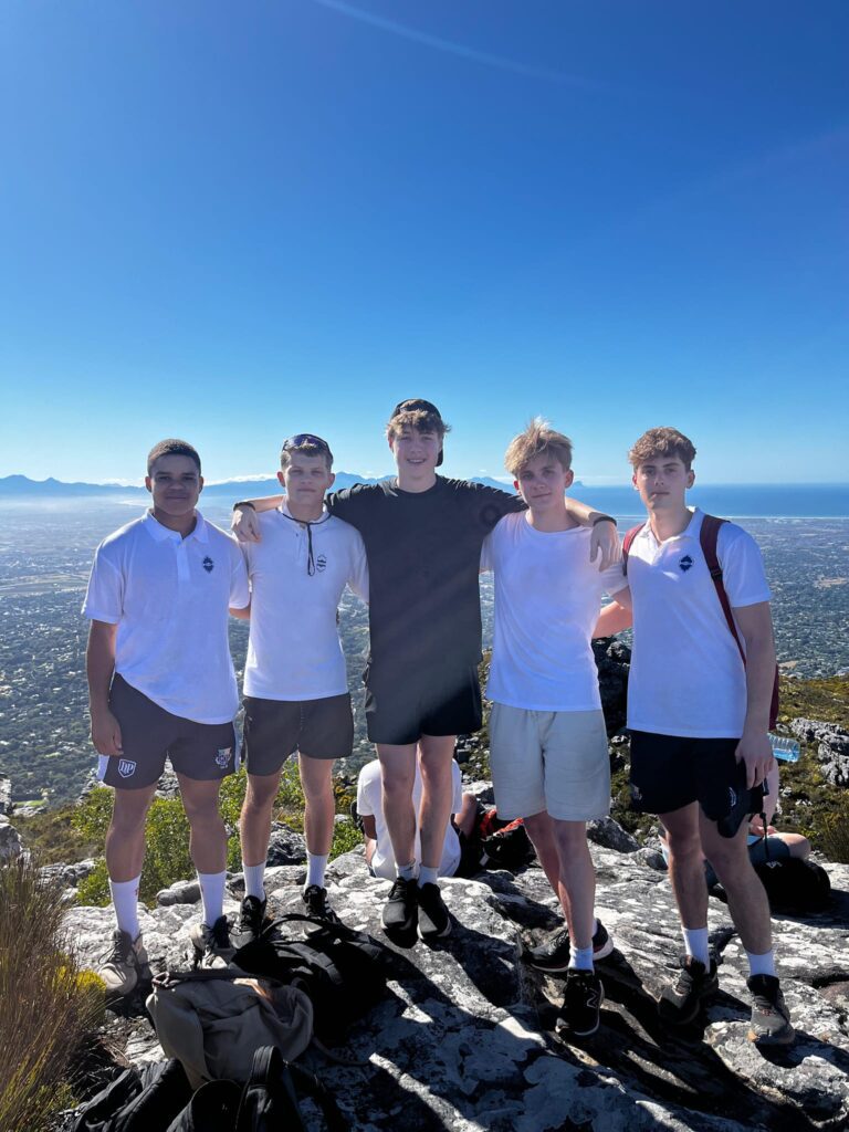 5 boys stand with their arms around each other with blue sky, mountains and city in the background.