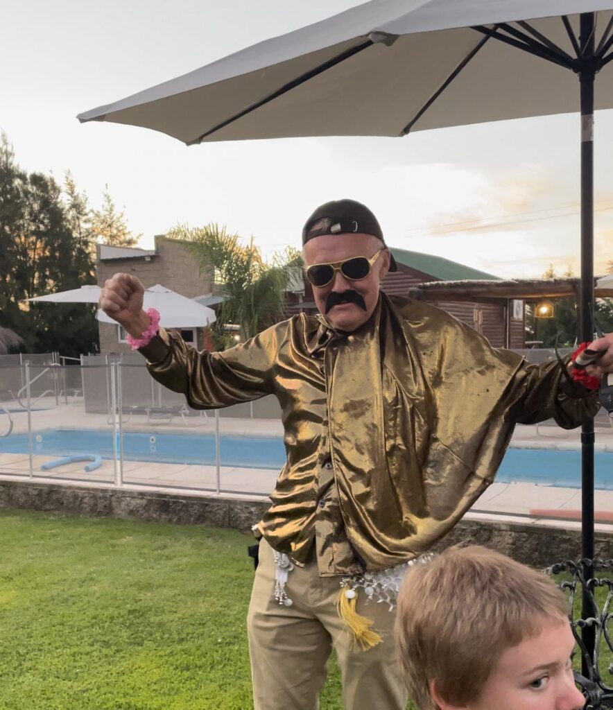 Older man stands with open arms, sunglasses and gold shirt, having fun.