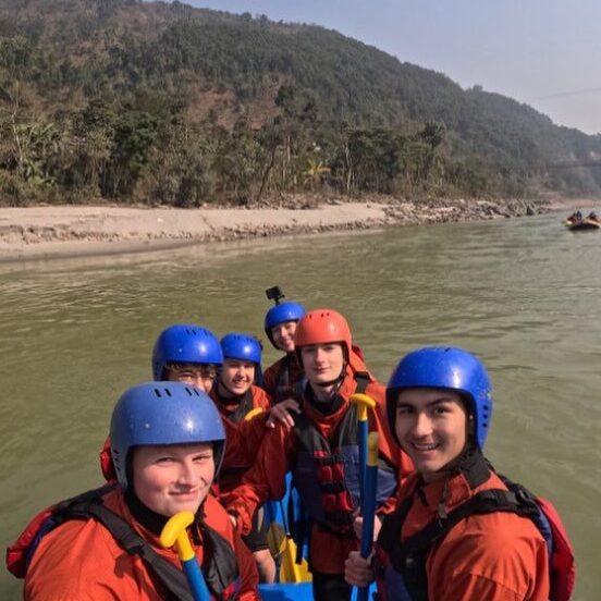 Students sit in a boat wearing blue helmets and orange suits