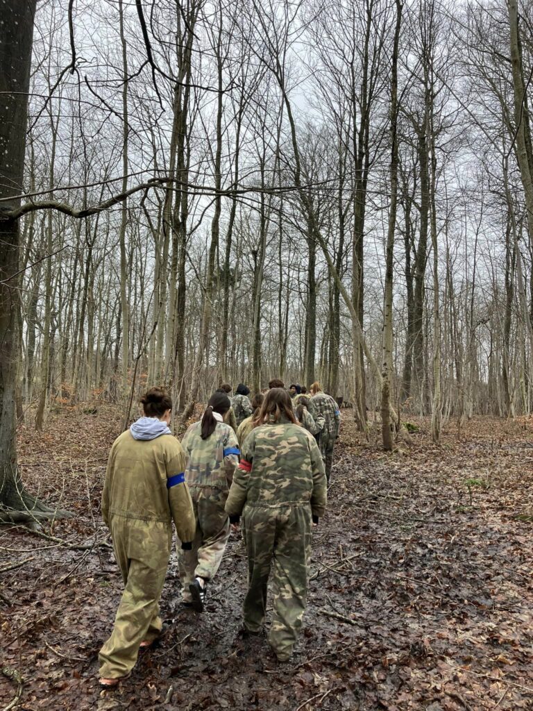 A group of students walk around in a forest wearing military green jumpsuits and playing paintball
