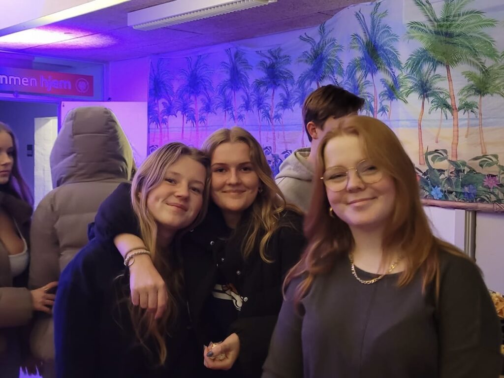 tre piger står og smiler mens to af den holder om hinanden