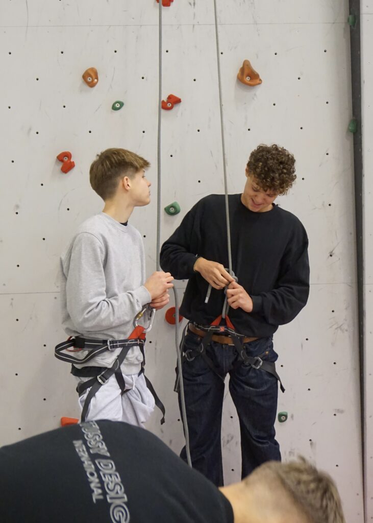 Two boys stand at a climbing wall and put on equipment