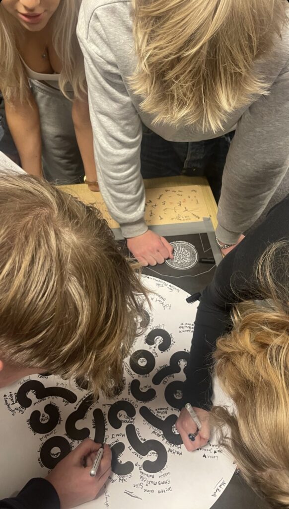 A group of students stand over a board with the Ranum efterskole college logo and write their names