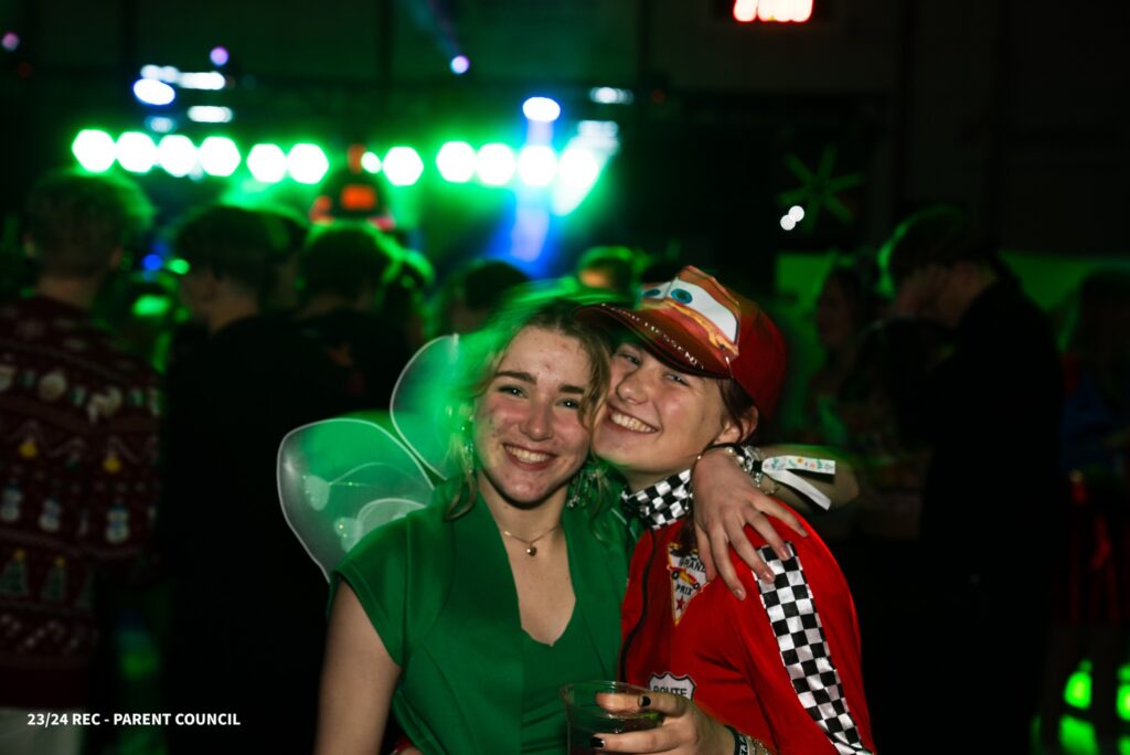 Two girls hold each other while dressed as a race car driver and a bellflower, with a colourful party in the background