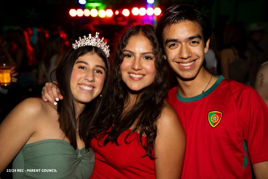 Two girls and a boy are holding each other and smiling, while a girl wears a crown on her head and there's a colourful party in the background