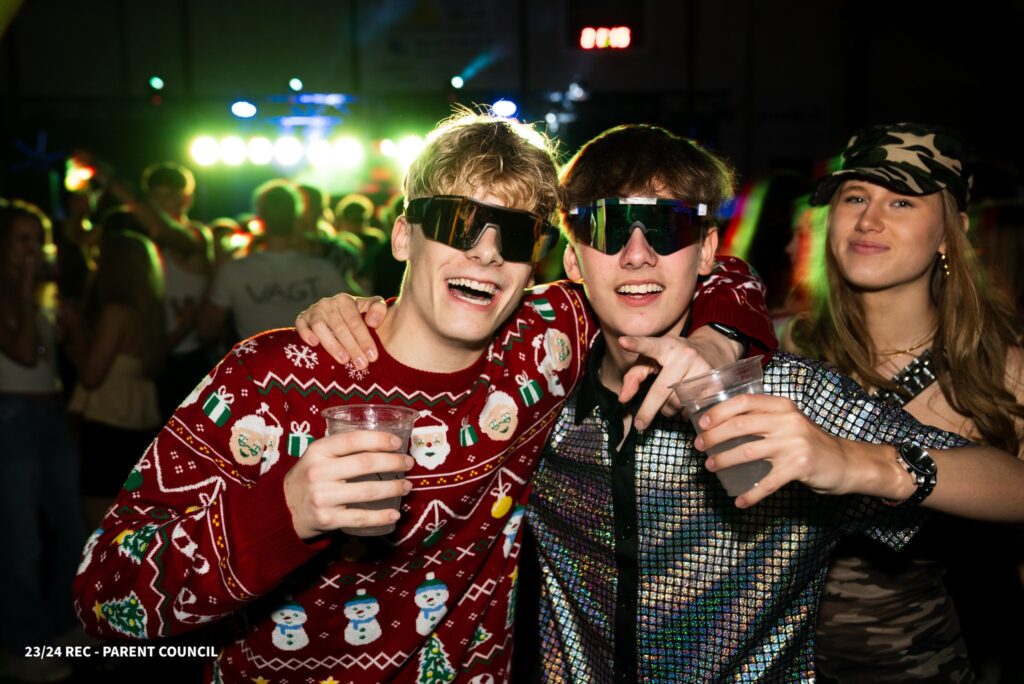 Two boys are holding each other and smiling while wearing sunglasses and holding a drink, with party and colour in the background