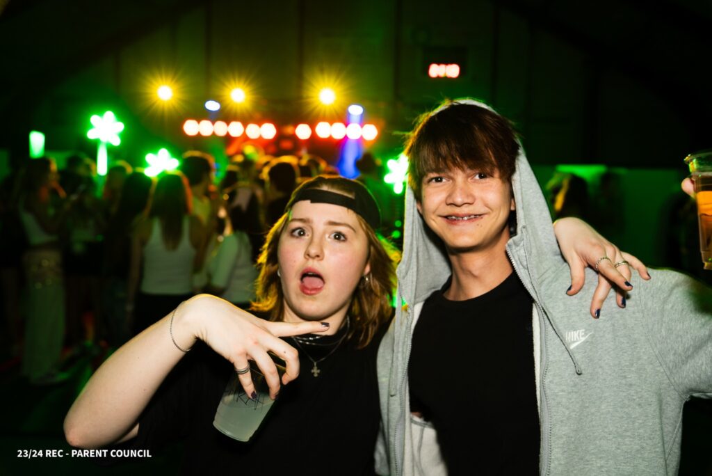 A girl and a boy are holding each other and smiling, and there's a colourful party in the background