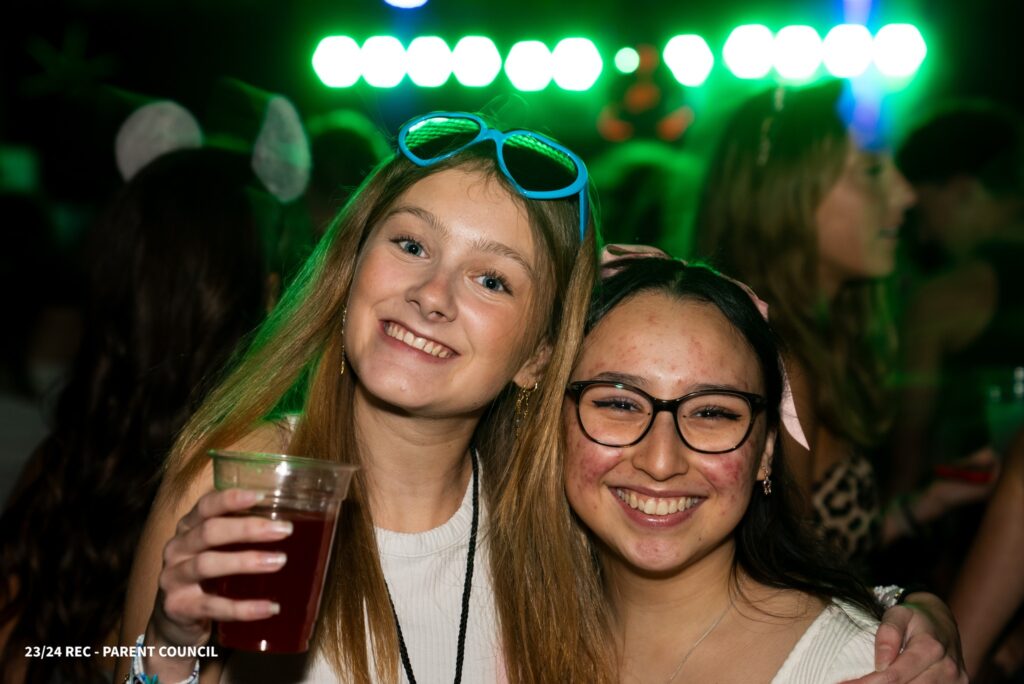 Two girls hold each other while smiling, and there's a colourful party in the background
